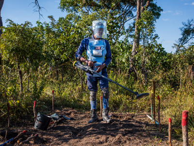 Male deminer wears PPE in a minefield whilst holding a metal detector