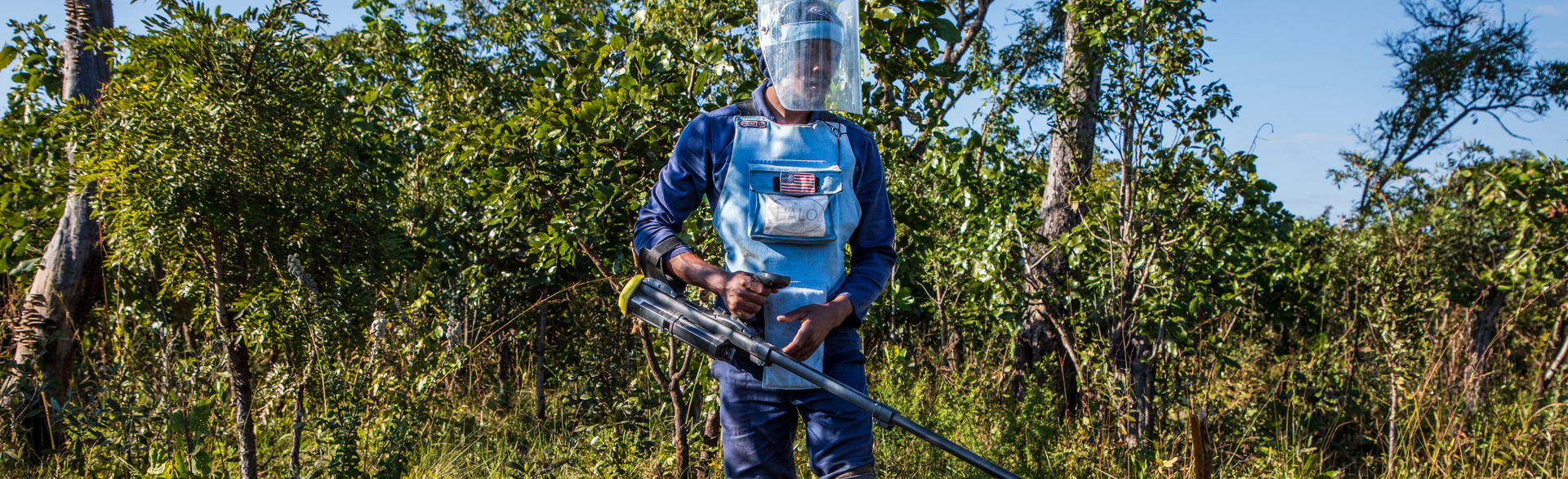 Male deminer wears PPE in a minefield whilst holding a metal detector