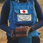A deminer wears protective gear with a Japan badge while holding a cube shaped block in her hands