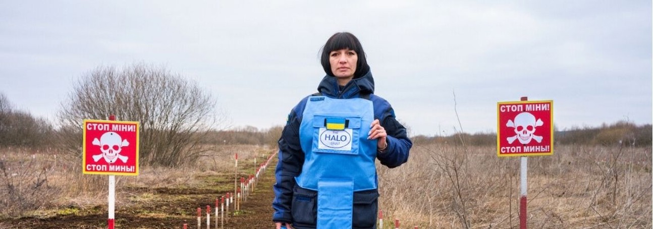 A woman in light blue HALO Ukraine demining gear stands between mine danger signs in a field.