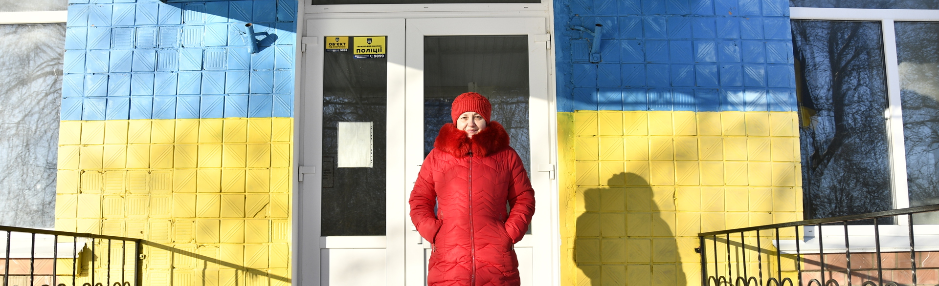 In a red coat and hat, Oksana stands in front of the school building's front painted in the Ukrainian flag