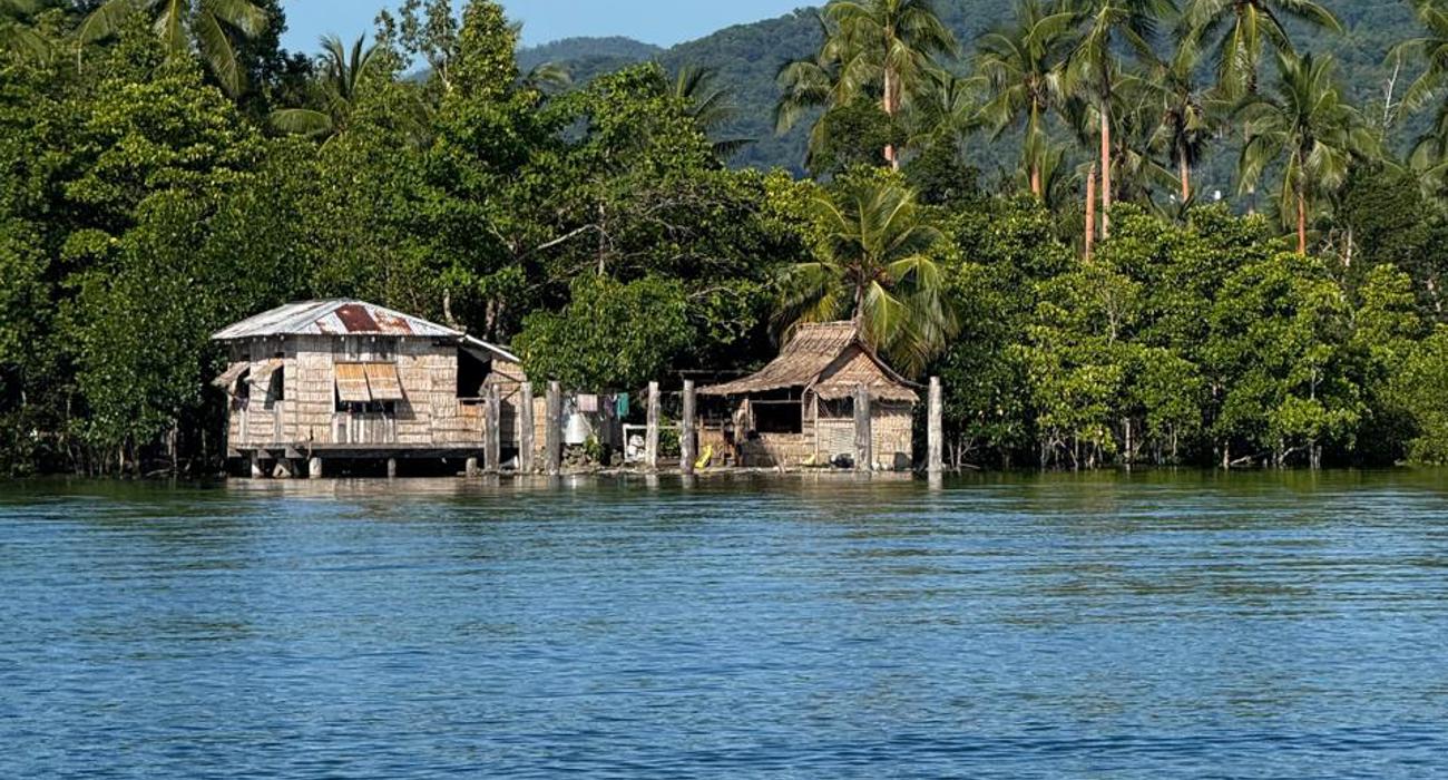 Two wooden houses on a blue lagoon, against a backdrop of trees and mountains.