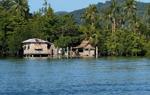 Two wooden houses on a blue lagoon, against a backdrop of trees and mountains.