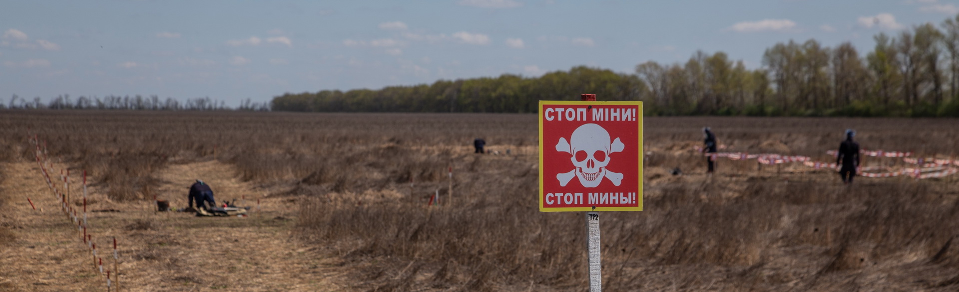 A sign says "Danger Mines" in Ukrainian while in the background 3 deminers manually clear a field