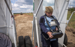 A woman in a HALO Trust vest operates a large remote control, controlling a large vehicle from a distance. 