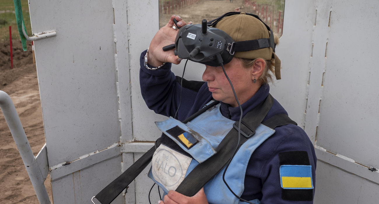 A woman in a HALO Trust vest operates a large remote control and looks into a pair of goggles.