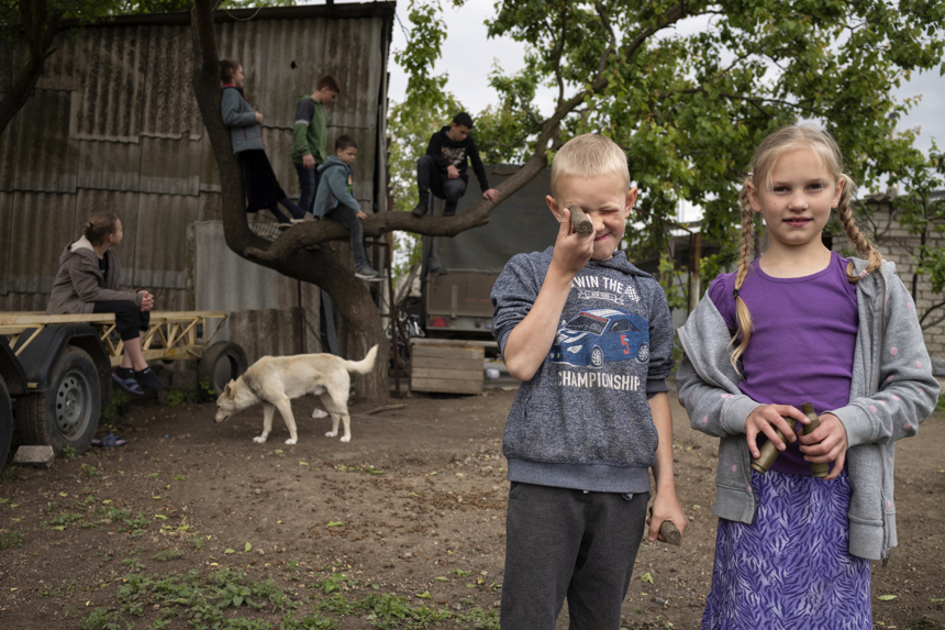 Two children face the camera, one looking through a small object, while other children play in a tree in the background.
