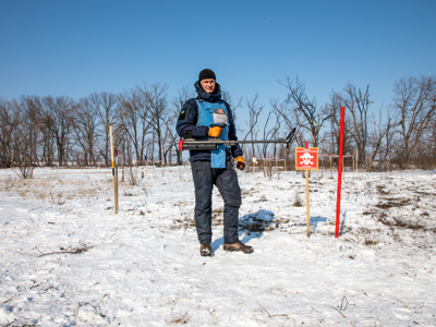 Deminer wearing a Ukrainian and an American flag holding a metal detector standing in a snow-covered minefield