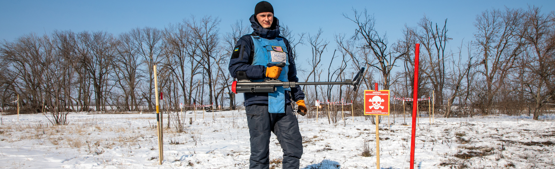 Deminer wearing a Ukrainian and an American flag holding a metal detector standing in a snow-covered minefield