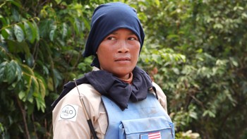 A female deminer stands in a blue vest with an American flag against a green background.