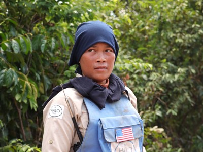 A female deminer stands in a blue vest with an American flag against a green background.