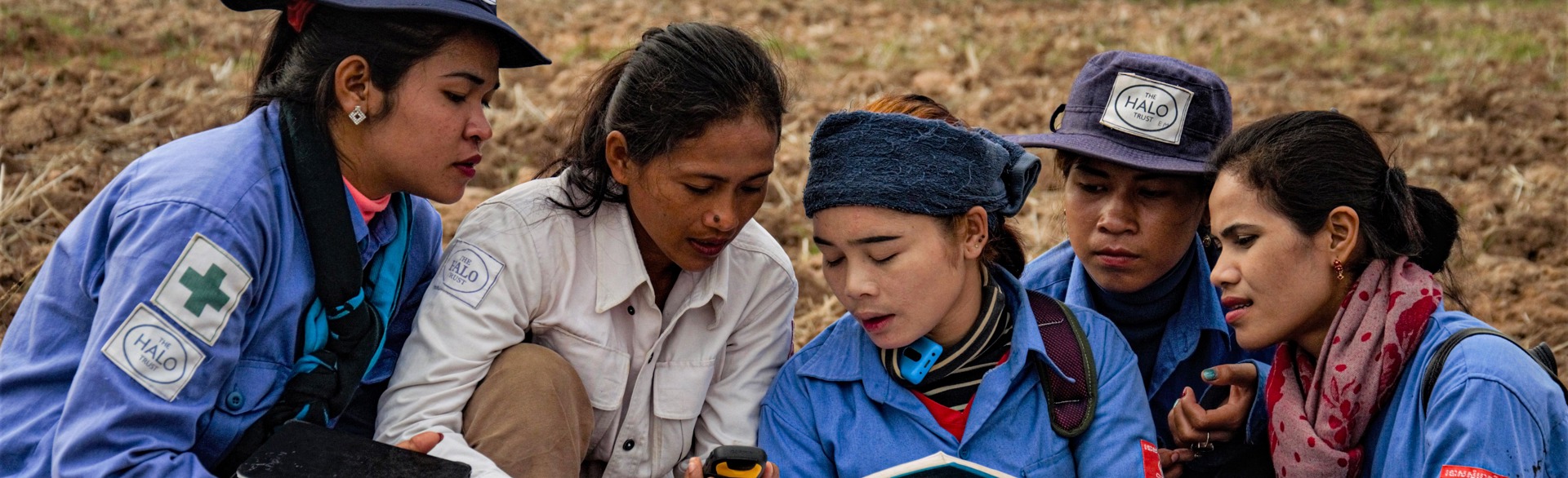 Five female deminers sit and look at a book held by the woman in the center.