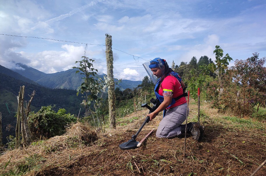 A woman in red and blue demining gear kneels at a mine site with mountains in the background