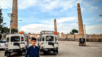 A boy stand in front of vehicles parked just outside the Herat Minarets