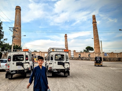 A boy stand in front of vehicles parked just outside the Herat Minarets