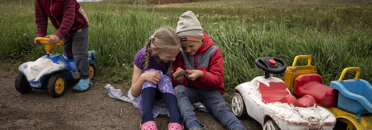 Two young children play together in a field. One boy rides a toy car to the left side.