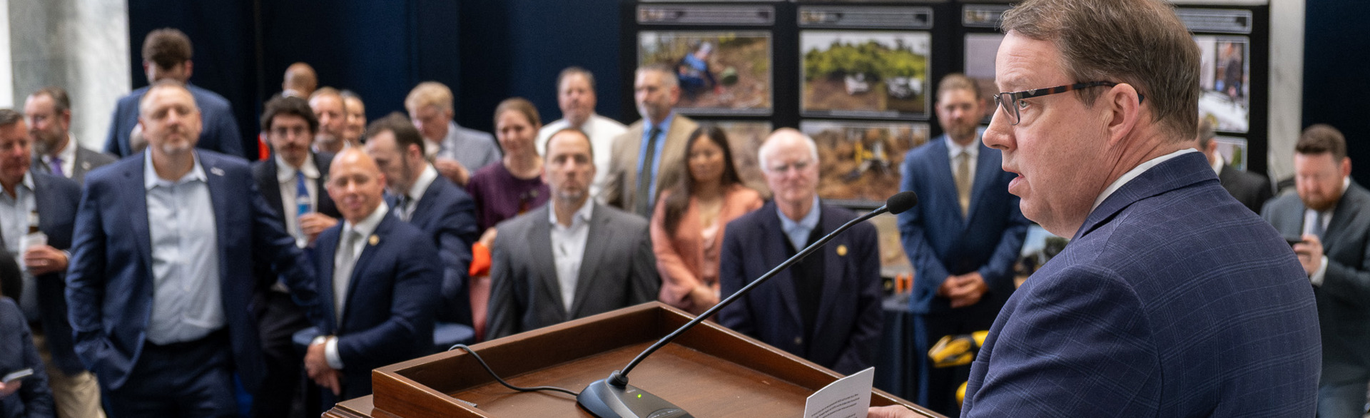 A man stands at the event podium speaking to a full room