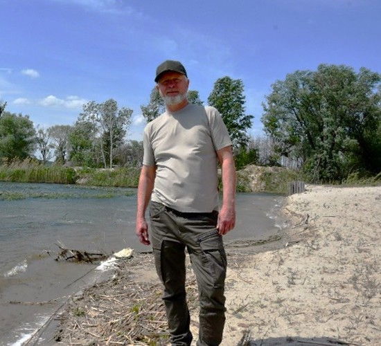 A man in a gray shirt and a cap stands on the shore of a lake.