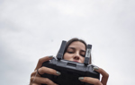 A woman in a blue jacket holds a controller while a drone flies overhead.