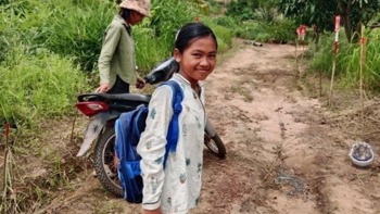 A young girl wearing a backpack stands in front of a path through high grasses marked by minefield signs on one side