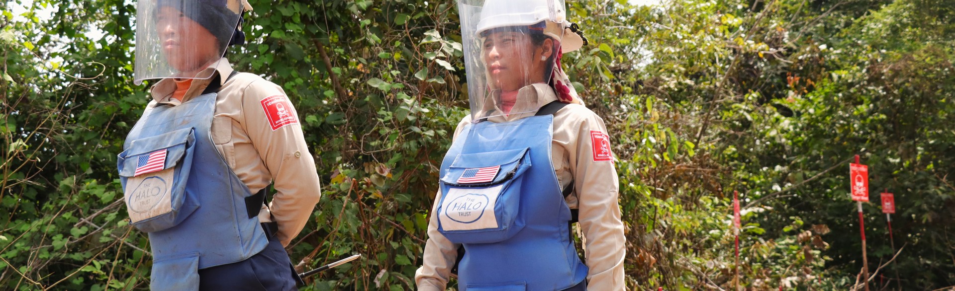 Two Cambodian deminers, wearing US Flags on their PPE vests, stand in a minefield