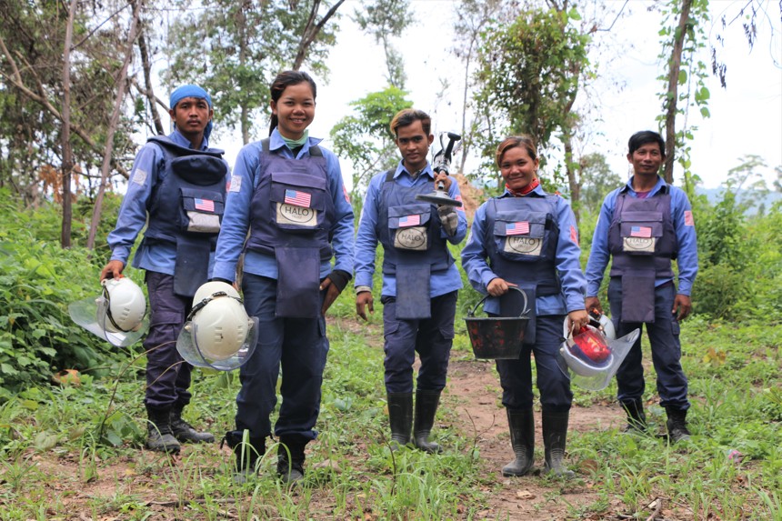 A team of HALO Trust deminers walk through a forest holding tools and helmets