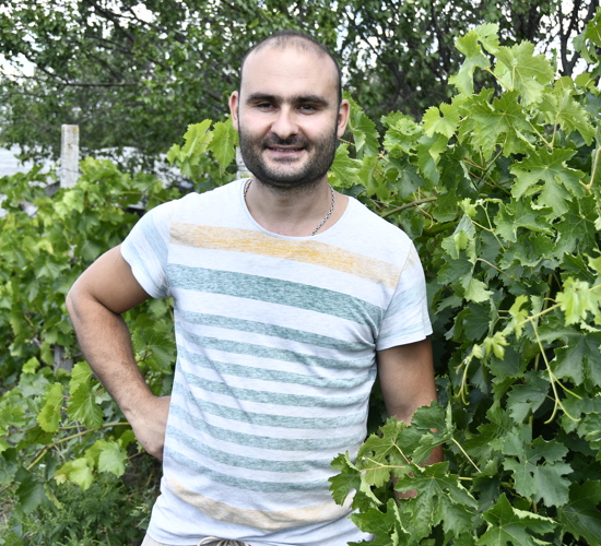 A man in a striped t-shirt stands smiling in front of a leafy bush.