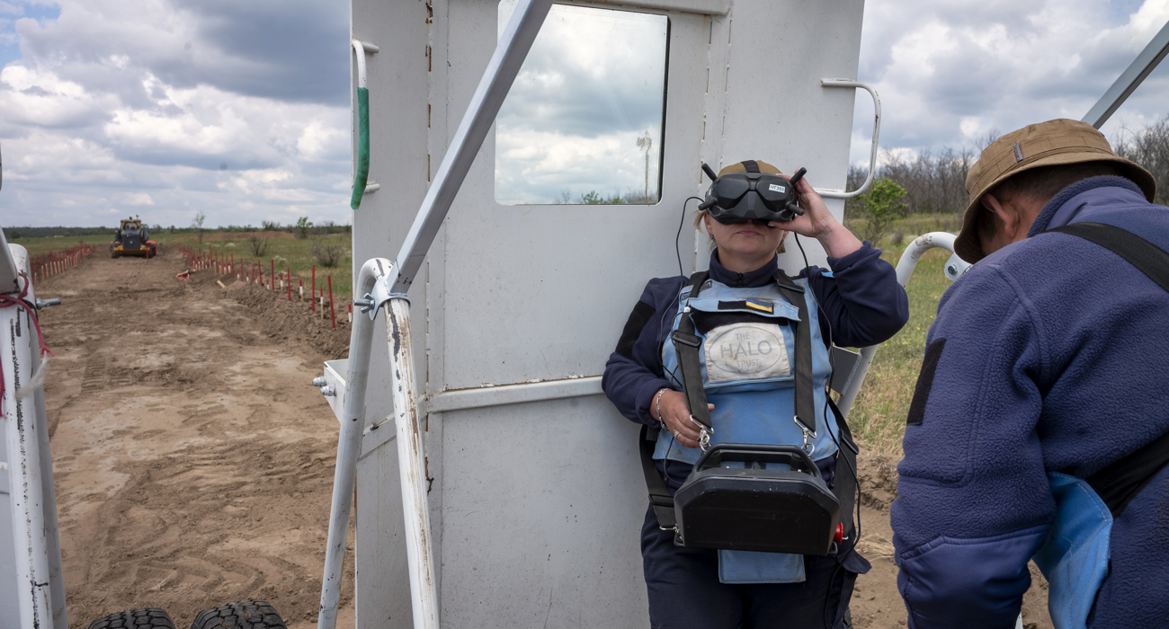 A woman in a HALO Trust vest operates a large remote control, controlling a large vehicle from a distance. 
