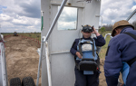 A woman in a HALO Trust vest operates a large remote control, controlling a large vehicle from a distance. 