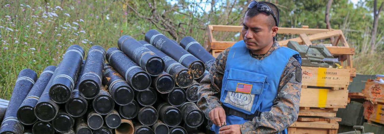A deminer stands in front of crates and cylinder containers outside
