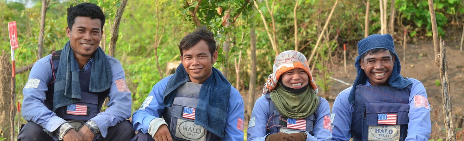 The demining team in Cambodia pose kneeling down and smile whilst wearing their PPE 