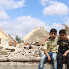 Two young boys sit in front of a destroyed house